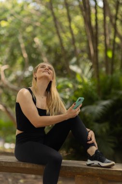 Gündüz vakti arka planda yeşil park olan akıllı telefon kullanan genç bir kadın. Yüksek kalite fotoğraf. Cep telefonu, teknoloji, şehir konsepti. Yüksek kalite fotoğraf