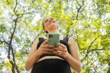 Gündüz vakti arka planda yeşil park olan akıllı telefon kullanan genç bir kadın. Yüksek kalite fotoğraf. Cep telefonu, teknoloji, şehir konsepti. Yüksek kalite fotoğraf