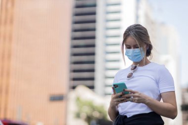 Gündüz vakti akıllı telefon kullanarak maske takan genç bir kadın. Arka planda şehir manzarası var. Salgın, covid 19, koronavirüs epiformasyonu. Güvenlik önlemleri. Yüksek kalite fotoğraf. Cep telefonu, teknoloji, şehir kavramı. 