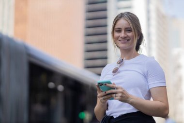Gündüz vakti arka planda şehir manzarası olan akıllı telefon kullanan genç bir kadın. Yüksek kalite fotoğraf. Cep telefonu, teknoloji, şehir kavramı. 