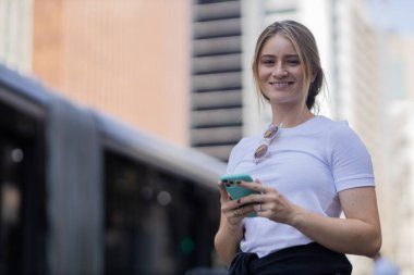 Gündüz vakti arka planda şehir manzarası olan akıllı telefon kullanan genç bir kadın. Yüksek kalite fotoğraf. Cep telefonu, teknoloji, şehir kavramı. 