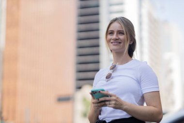 Gündüz vakti arka planda şehir manzarası olan akıllı telefon kullanan genç bir kadın. Yüksek kalite fotoğraf. Cep telefonu, teknoloji, şehir kavramı. 