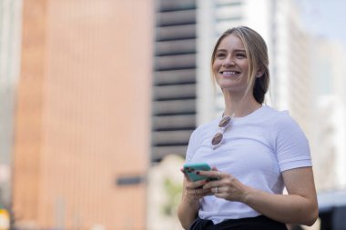 Gündüz vakti arka planda şehir manzarası olan akıllı telefon kullanan genç bir kadın. Yüksek kalite fotoğraf. Cep telefonu, teknoloji, şehir kavramı. 