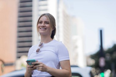 Gündüz vakti arka planda şehir manzarası olan akıllı telefon kullanan genç bir kadın. Yüksek kalite fotoğraf. Cep telefonu, teknoloji, şehir kavramı. 