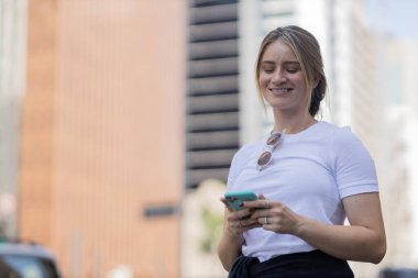 Gündüz vakti arka planda şehir manzarası olan akıllı telefon kullanan genç bir kadın. Yüksek kalite fotoğraf. Cep telefonu, teknoloji, şehir kavramı. 