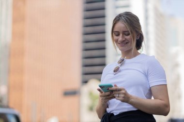 Gündüz vakti arka planda şehir manzarası olan akıllı telefon kullanan genç bir kadın. Yüksek kalite fotoğraf. Cep telefonu, teknoloji, şehir kavramı. 