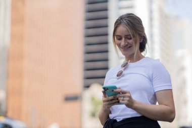 Gündüz vakti arka planda şehir manzarası olan akıllı telefon kullanan genç bir kadın. Yüksek kalite fotoğraf. Cep telefonu, teknoloji, şehir kavramı. 
