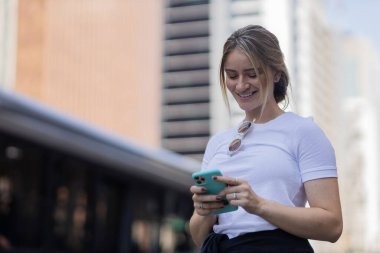 Gündüz vakti arka planda şehir manzarası olan akıllı telefon kullanan genç bir kadın. Yüksek kalite fotoğraf. Cep telefonu, teknoloji, şehir kavramı. 