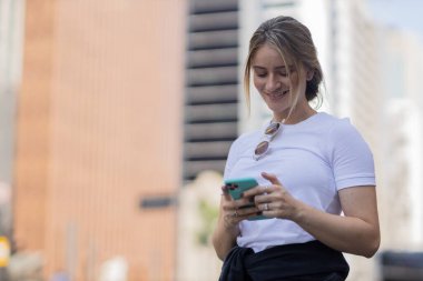 Gündüz vakti arka planda şehir manzarası olan akıllı telefon kullanan genç bir kadın. Yüksek kalite fotoğraf. Cep telefonu, teknoloji, şehir kavramı. 