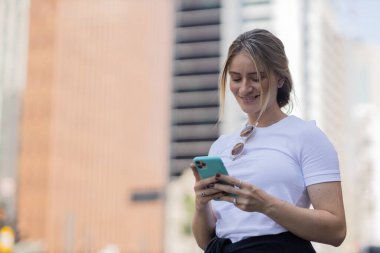 Gündüz vakti arka planda şehir manzarası olan akıllı telefon kullanan genç bir kadın. Yüksek kalite fotoğraf. Cep telefonu, teknoloji, şehir kavramı. 