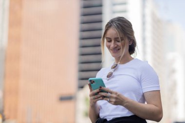 Gündüz vakti arka planda şehir manzarası olan akıllı telefon kullanan genç bir kadın. Yüksek kalite fotoğraf. Cep telefonu, teknoloji, şehir kavramı. 