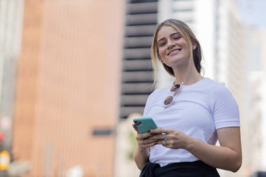 Gündüz vakti arka planda şehir manzarası olan akıllı telefon kullanan genç bir kadın. Yüksek kalite fotoğraf. Cep telefonu, teknoloji, şehir kavramı. 