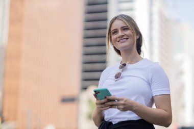 Gündüz vakti arka planda şehir manzarası olan akıllı telefon kullanan genç bir kadın. Yüksek kalite fotoğraf. Cep telefonu, teknoloji, şehir kavramı. 