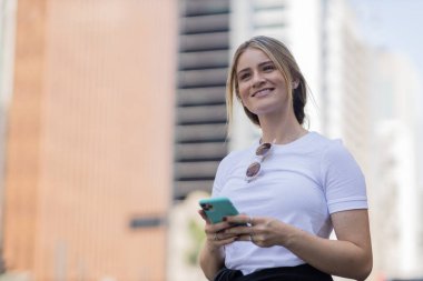 Gündüz vakti arka planda şehir manzarası olan akıllı telefon kullanan genç bir kadın. Yüksek kalite fotoğraf. Cep telefonu, teknoloji, şehir kavramı. 
