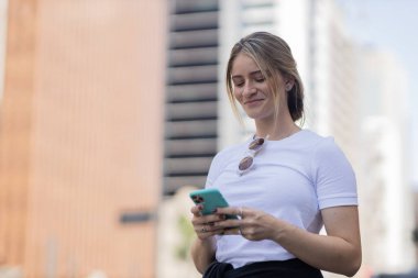 Gündüz vakti arka planda şehir manzarası olan akıllı telefon kullanan genç bir kadın. Yüksek kalite fotoğraf. Cep telefonu, teknoloji, şehir kavramı. 