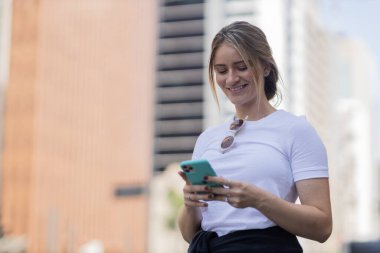 Gündüz vakti arka planda şehir manzarası olan akıllı telefon kullanan genç bir kadın. Yüksek kalite fotoğraf. Cep telefonu, teknoloji, şehir kavramı. 