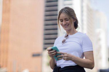 Gündüz vakti arka planda şehir manzarası olan akıllı telefon kullanan genç bir kadın. Yüksek kalite fotoğraf. Cep telefonu, teknoloji, şehir kavramı. 