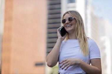 Gündüz vakti arka planda şehir manzarası olan akıllı telefon kullanan genç bir kadın. Yüksek kalite fotoğraf. Cep telefonu, teknoloji, şehir kavramı. 
