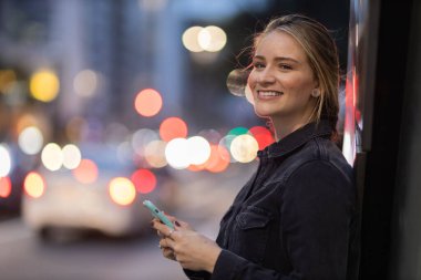 Geceleri arka planda şehir manzarası olan akıllı telefon kullanan genç bir kadın. Yüksek kalite fotoğraf. Fotoğraf. Cep telefonu, teknoloji, şehir kavramı. 
