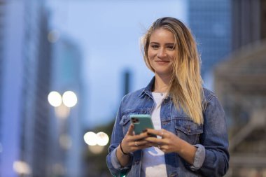 Geceleri arka planda şehir manzarası olan akıllı telefon kullanan genç bir kadın. Yüksek kalite fotoğraf. Fotoğraf. Cep telefonu, teknoloji, şehir kavramı. 