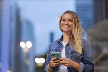 Geceleri arka planda şehir manzarası olan akıllı telefon kullanan genç bir kadın. Yüksek kalite fotoğraf. Fotoğraf. Cep telefonu, teknoloji, şehir kavramı. 