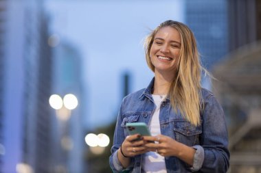 Geceleri arka planda şehir manzarası olan akıllı telefon kullanan genç bir kadın. Yüksek kalite fotoğraf. Fotoğraf. Cep telefonu, teknoloji, şehir kavramı. 