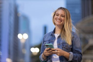 Geceleri arka planda şehir manzarası olan akıllı telefon kullanan genç bir kadın. Yüksek kalite fotoğraf. Fotoğraf. Cep telefonu, teknoloji, şehir kavramı. 