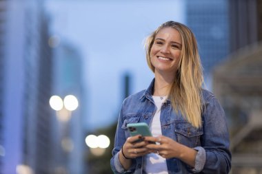 Geceleri arka planda şehir manzarası olan akıllı telefon kullanan genç bir kadın. Yüksek kalite fotoğraf. Fotoğraf. Cep telefonu, teknoloji, şehir kavramı. 