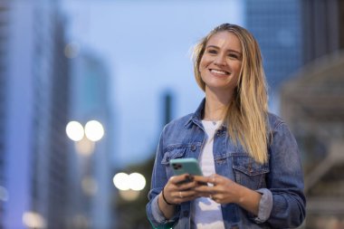 Geceleri arka planda şehir manzarası olan akıllı telefon kullanan genç bir kadın. Yüksek kalite fotoğraf. Fotoğraf. Cep telefonu, teknoloji, şehir kavramı. 