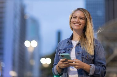 Geceleri arka planda şehir manzarası olan akıllı telefon kullanan genç bir kadın. Yüksek kalite fotoğraf. Fotoğraf. Cep telefonu, teknoloji, şehir kavramı. 