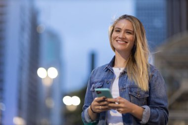 Geceleri arka planda şehir manzarası olan akıllı telefon kullanan genç bir kadın. Yüksek kalite fotoğraf. Fotoğraf. Cep telefonu, teknoloji, şehir kavramı. 
