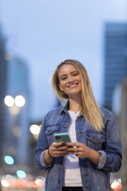 Geceleri arka planda şehir manzarası olan akıllı telefon kullanan genç bir kadın. Yüksek kalite fotoğraf. Fotoğraf. Cep telefonu, teknoloji, şehir kavramı. 