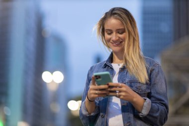 Geceleri arka planda şehir manzarası olan akıllı telefon kullanan genç bir kadın. Yüksek kalite fotoğraf. Fotoğraf. Cep telefonu, teknoloji, şehir kavramı. 