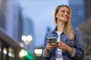 Geceleri arka planda şehir manzarası olan akıllı telefon kullanan genç bir kadın. Yüksek kalite fotoğraf. Fotoğraf. Cep telefonu, teknoloji, şehir kavramı. 
