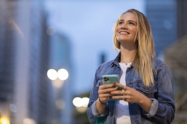 Geceleri arka planda şehir manzarası olan akıllı telefon kullanan genç bir kadın. Yüksek kalite fotoğraf. Fotoğraf. Cep telefonu, teknoloji, şehir kavramı. 