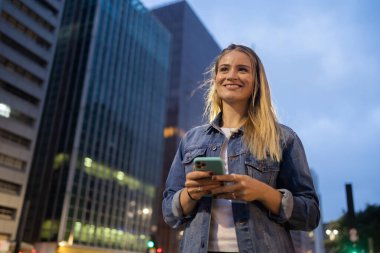 Geceleri arka planda şehir manzarası olan akıllı telefon kullanan genç bir kadın. Yüksek kalite fotoğraf. Fotoğraf. Cep telefonu, teknoloji, şehir kavramı. 