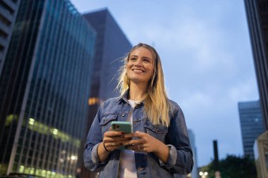 Geceleri arka planda şehir manzarası olan akıllı telefon kullanan genç bir kadın. Yüksek kalite fotoğraf. Fotoğraf. Cep telefonu, teknoloji, şehir kavramı. 