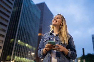 Geceleri arka planda şehir manzarası olan akıllı telefon kullanan genç bir kadın. Yüksek kalite fotoğraf. Fotoğraf. Cep telefonu, teknoloji, şehir kavramı. 