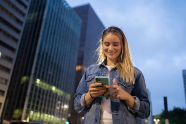 Geceleri arka planda şehir manzarası olan akıllı telefon kullanan genç bir kadın. Yüksek kalite fotoğraf. Fotoğraf. Cep telefonu, teknoloji, şehir kavramı. 