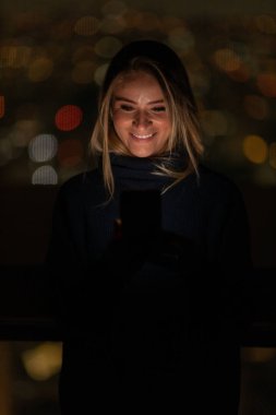 Geceleri arka planda şehir manzarası olan akıllı telefon kullanan genç bir kadın. Yüksek kalite fotoğraf. Cep telefonu, teknoloji, konsept. Yüksek kalite fotoğraf