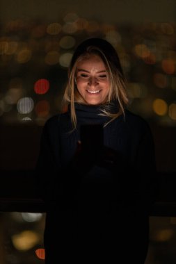 Geceleri arka planda şehir manzarası olan akıllı telefon kullanan genç bir kadın. Yüksek kalite fotoğraf. Cep telefonu, teknoloji, konsept. Yüksek kalite fotoğraf