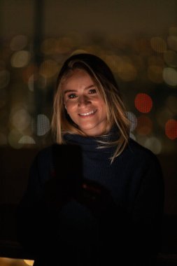 Geceleri arka planda şehir manzarası olan akıllı telefon kullanan genç bir kadın. Yüksek kalite fotoğraf. Cep telefonu, teknoloji, konsept. Yüksek kalite fotoğraf