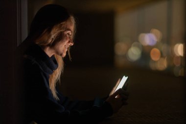 Geceleri arka planda şehir manzarası olan akıllı telefon kullanan genç bir kadın. Yüksek kalite fotoğraf. Cep telefonu, teknoloji, konsept. Yüksek kalite fotoğraf