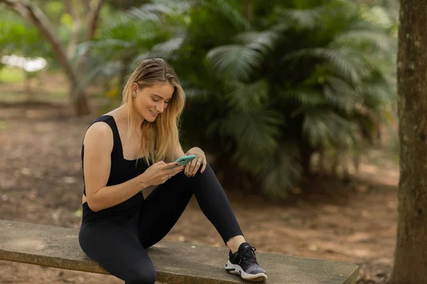 Gündüz vakti arka planda yeşil park olan akıllı telefon kullanan genç bir kadın. Yüksek kalite fotoğraf. Cep telefonu, teknoloji, şehir konsepti. Yüksek kalite fotoğraf