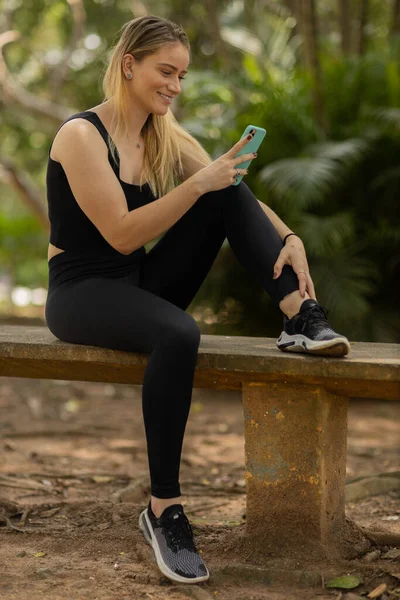 Gündüz vakti arka planda yeşil park olan akıllı telefon kullanan genç bir kadın. Yüksek kalite fotoğraf. Cep telefonu, teknoloji, şehir konsepti. Yüksek kalite fotoğraf