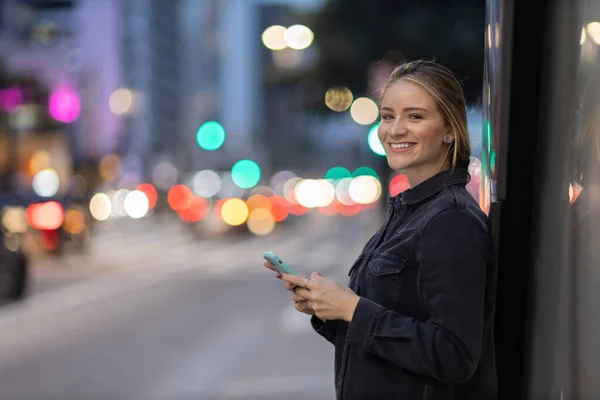 Geceleri arka planda şehir manzarası olan akıllı telefon kullanan genç bir kadın. Yüksek kalite fotoğraf. Fotoğraf. Cep telefonu, teknoloji, şehir kavramı. 