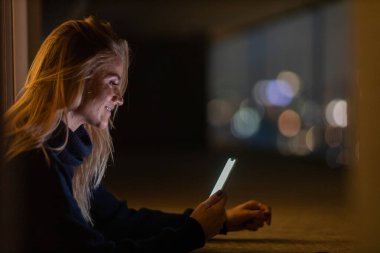 Geceleri arka planda şehir manzarası olan akıllı telefon kullanan genç bir kadın. Yüksek kalite fotoğraf. Cep telefonu, teknoloji, konsept. Yüksek kalite fotoğraf