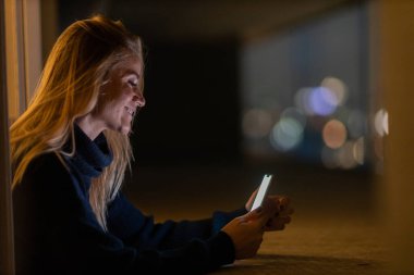 Geceleri arka planda şehir manzarası olan akıllı telefon kullanan genç bir kadın. Yüksek kalite fotoğraf. Cep telefonu, teknoloji, konsept. Yüksek kalite fotoğraf