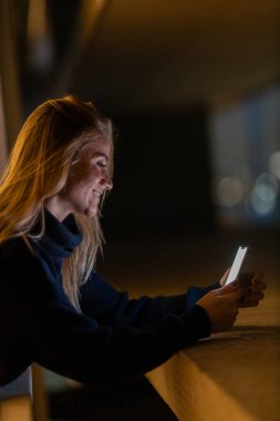 Geceleri arka planda şehir manzarası olan akıllı telefon kullanan genç bir kadın. Yüksek kalite fotoğraf. Cep telefonu, teknoloji, konsept. Yüksek kalite fotoğraf