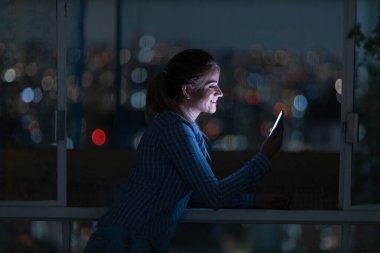 Geceleri arka planda şehir manzarası olan akıllı telefon kullanan genç bir kadın. Yüksek kalite fotoğraf. Cep telefonu, teknoloji, konsept. Yüksek kalite fotoğraf